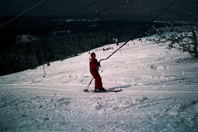 Mid-century skier in red ensemble descends snowy slope under artificial lighting, likely a ski lift station. Snow-covered ter...