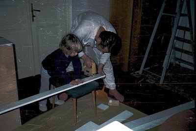Two children in 1970s-style clothing repair a wooden beam indoors, supported by a ladder and plastic sheeting. The setting ap...