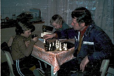 Three individuals engage in a chess game at a round table draped in patterned fabric, likely mid-1970s. The adult wears a str...
