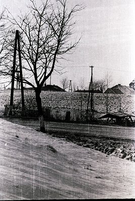Mid-20th century rural street scene with stone wall and bare trees. Snow-covered road suggests winter, likely Eastern Europe....