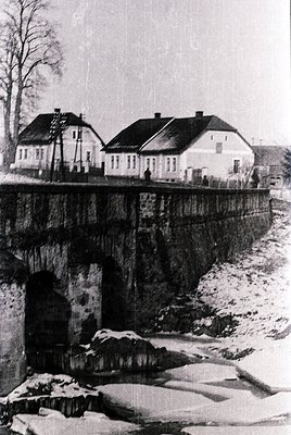Mid-20th century rural bridge spanning a shallow, snow-covered stream. Two-story brick houses with pitched roofs and chimneys...