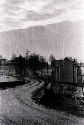 Mid-20th century street scene with curved roadway and industrial-era buildings. Leafless trees line a paved curve flanked by ...