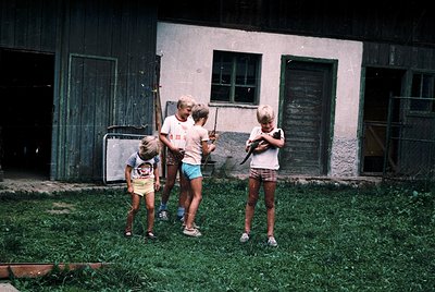 Four children in 1970s-style clothing play with baseball bats in front of a weathered, green-painted wooden building. The adu...