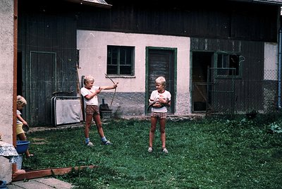Two children playing with a broomstick in a courtyard, 1960s-1970s. Rustic brick building with faded paint, green window fram...