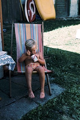 Mid-20th-century beachside scene: young child in red-and-white swim diaper sits on a striped deck chair, eating an ice cream ...