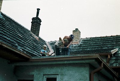 Two adults and a child repairing a tiled roof in a rural setting, likely Eastern Europe, mid-20th century. The man in the cen...