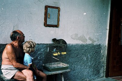 Vintage interior shot of elderly man and child observing a birdcage with a canary. Plain walls, simple wooden door, and worn ...