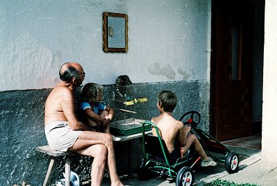 Three generations play ping-pong in a sunlit courtyard, framed by weathered plaster walls and a wooden door. The elderly man ...