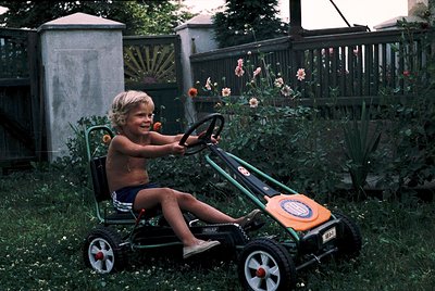 Young child in 1970s-style swim trunks operates a vintage toy go-kart in a lush backyard. Concrete block wall and wooden fenc...