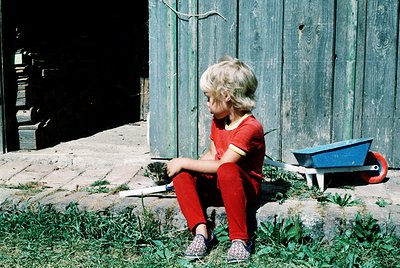 A young child in red clothing sits on grass beside a weathered wooden fence, holding a small wooden object. A blue wheelbarro...