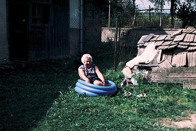 Vintage photo of elderly woman seated in a blue inflatable pool in a rural backyard. Wooden shed and rustic fence with overgr...
