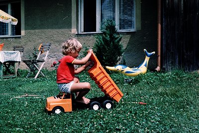 Child kneeling on grass, pushing a vintage orange toy dump truck in a backyard. Yellow airplane toy and wooden ladder in back...