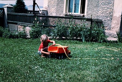 Child in vintage orange toy tractor on lush green lawn, mid-20th century residential setting. Wrought-iron fence and stone ho...