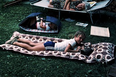 Vintage outdoor camping scene with three generations lounging on patterned mattresses on grass. Adults in 1970s-style clothin...