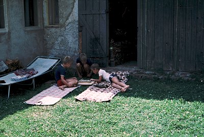 Three children play on grass near a rustic stone building with wooden doors, seated on patterned blankets. Mid-20th century r...