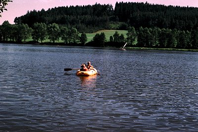 Two individuals paddle a bright orange inflatable raft on calm waters, framed by dense forested hills and a distant sailboat....