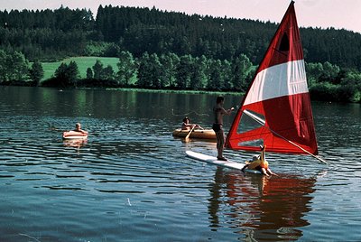 Windsurfer in red/white sail navigates serene lake surrounded by dense forest. Two individuals in watercraft—one on a paddleb...