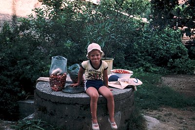 Child poses on concrete planter beside lush greenery, surrounded by jars of cherries and a tray of fresh produce. Mid-20th ce...