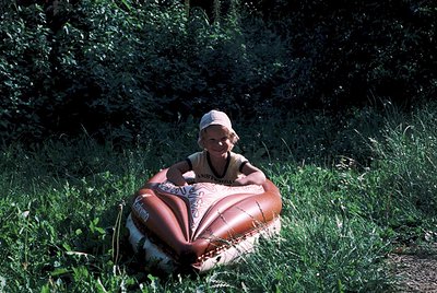 Child sitting on a vintage inflatable heart-shaped pool toy in a grassy, wooded area. Sunlight filters through dense foliage,...