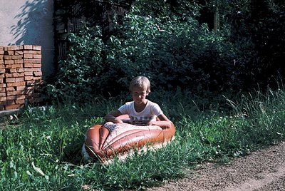 Child sits on an orange inner tube in a grassy backyard, mid-1970s. Brick wall and lush greenery frame the scene. Casual summ...