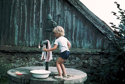 Child stands on stone pedestal washing clothes in vintage outdoor laundry sink beside weathered wooden barn, 1960s-70s rural ...