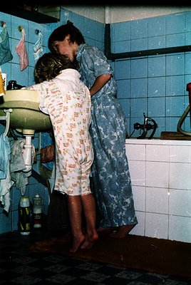 Intimate moment in a retro bathroom with blue-tiled walls, featuring two individuals in patterned pajamas embracing near a si...