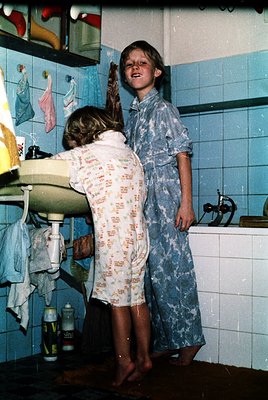 Two children in matching floral pajamas play in a retro bathroom with blue tiles, 1970s style. One stands on a pedestal sink,...
