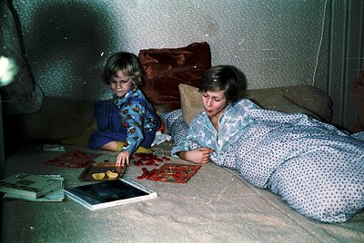 Two children in 1970s-style pajamas play on a patterned sofa, surrounded by vintage board games and a tray of food. The boy s...