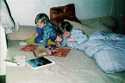 Two children in 1980s-style pajamas play on a patterned bedspread, surrounded by scattered magazines and a small photo album....