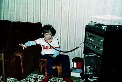 Vintage 1970s-80s interior shot: child wearing headphones connected to a bulky ABBA Greatest Hits cassette deck, seated on a ...