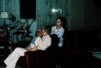 Vintage indoor portrait of three generations in a dimly lit living room, likely 1970s. Elderly man in glasses and patterned s...