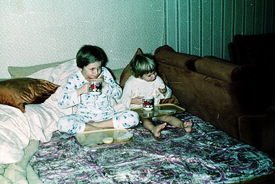 Two children in 1970s-style pajamas sit on a floral-patterned bed, sharing a snack. The boy holds a red-and-white cup, the gi...
