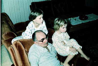 Vintage indoor portrait of a man and two children in matching floral pajamas on a brown leather couch, 1970s. The man wears g...