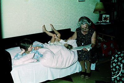 Vintage indoor scene featuring two children lying on a bed with a woman in a floral dress adjusting a child’s hair. Mid-20th ...