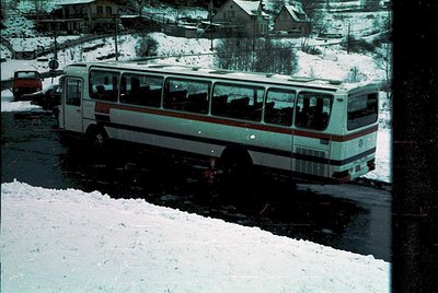 Mid-1970s white-and-red coach bus navigating snow-covered road in rural European village. Snow-laden trees and residential ho...