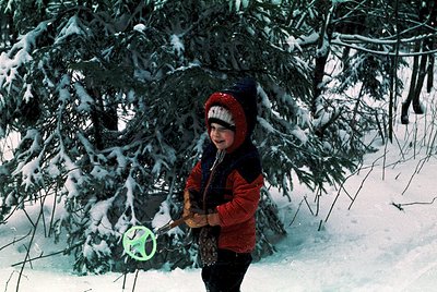 Child in winter forest holding neon green frisbee, bundled in red knit hat and red/orange jacket. Snow-covered evergreens fra...