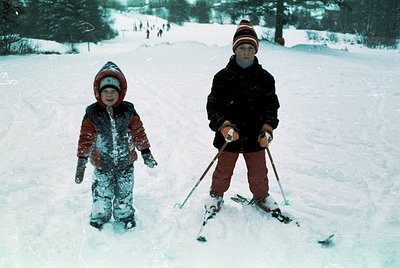 Two children cross-country skiing on snow-covered trails, mid-20th century winter attire. The younger wears a snowsuit with a...