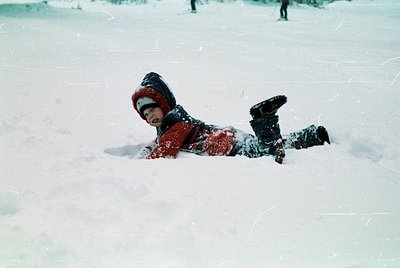 Child in vintage winter gear—red snowsuit, black boots, and hood—playing in snow, mid-roll. Blurry background suggests motion...