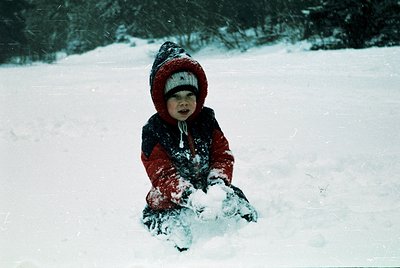 Child in winter attire builds snow pile in open snowy field. Red hooded jacket, dark pants, and mittens contrast against whit...