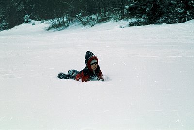 Child in vintage winter gear—red beanie and jacket—lying in snow-covered terrain, surrounded by forested hills. Likely mid-20...