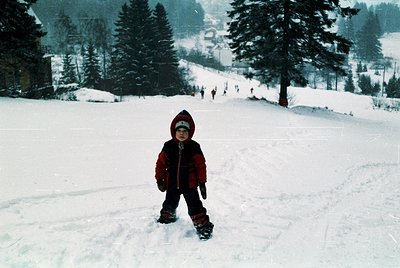 Child in vintage ski gear poses on snow-covered slope, surrounded by dense pine forest. Snowy alpine setting with distant ski...
