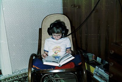 Vintage indoor shot of a child engrossed in a book, seated in a wicker armchair with headphones. Mid-20th century home settin...