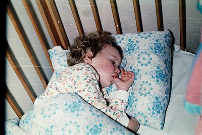 Vintage photograph of a young child peacefully sleeping in a crib, dressed in floral-patterned pajamas with blue and white fl...