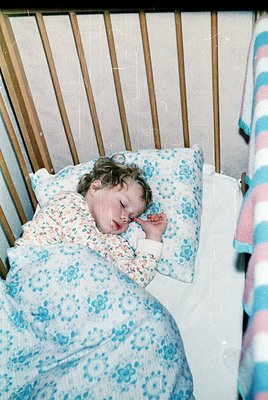 Vintage hospital crib with metal slats, featuring a child sleeping in a floral-patterned hospital gown and blue/polka-dot pil...