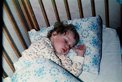 Vintage-style photo of an infant peacefully sleeping in a crib, dressed in a floral-patterned onesie with blue and white flor...