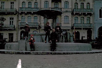 Vintage urban scene featuring a group of four children posing on a decorative fountain in a European city square. Architectur...