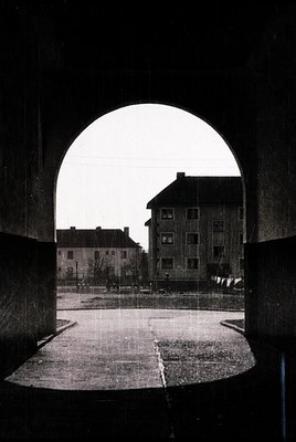 Black-and-white urban shot through a large, arched concrete tunnel entrance, revealing a residential courtyard. Multi-story a...