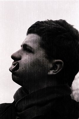 Portrait of a man in profile, captured mid-speech with rain streaks obscuring his face. Dark, textured jacket suggests cold w...