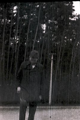 Vintage black-and-white shot of a man in a sweater and trousers standing under a rain curtain in a forest. Vertical wooden sl...