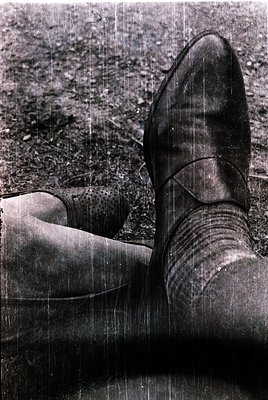 Vintage black-and-white shot of a person’s legs in worn leather boots, sitting on rough gravel. Distressed texture suggests m...
